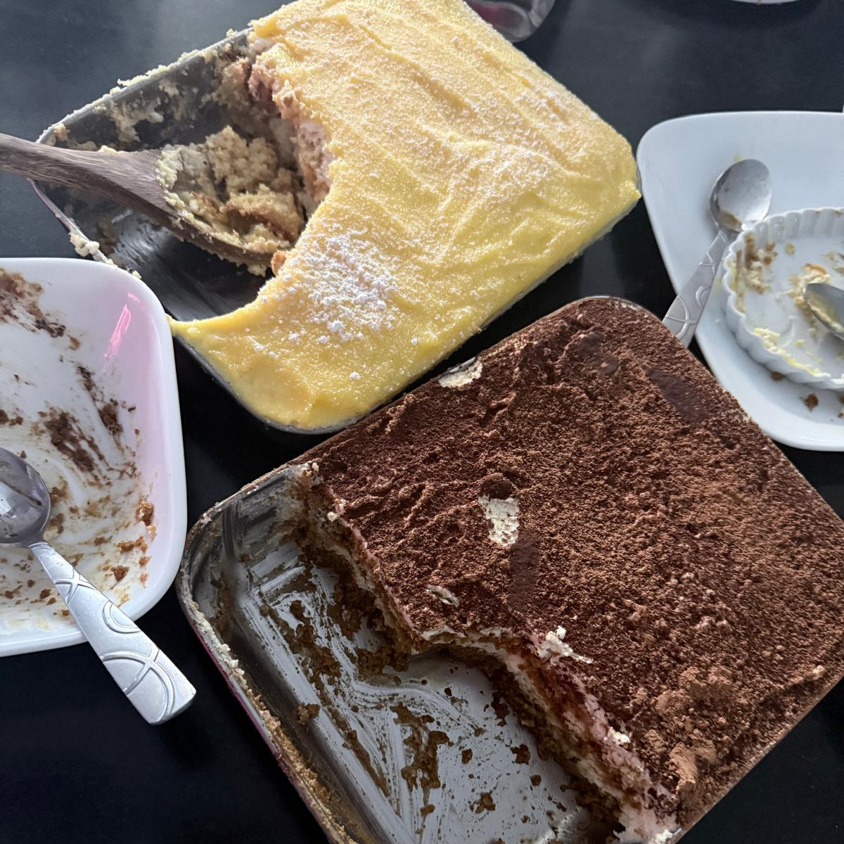 Two slices of cake in metal pans on a dark surface with utensils around.