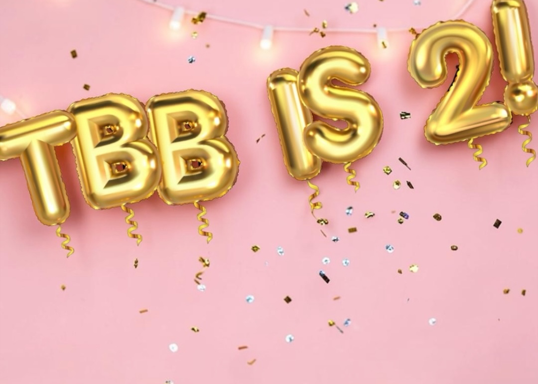 Gold foil balloon letters spelling 'TBB IS 2!' arranged as a banner on a pink background with string lights and confetti.